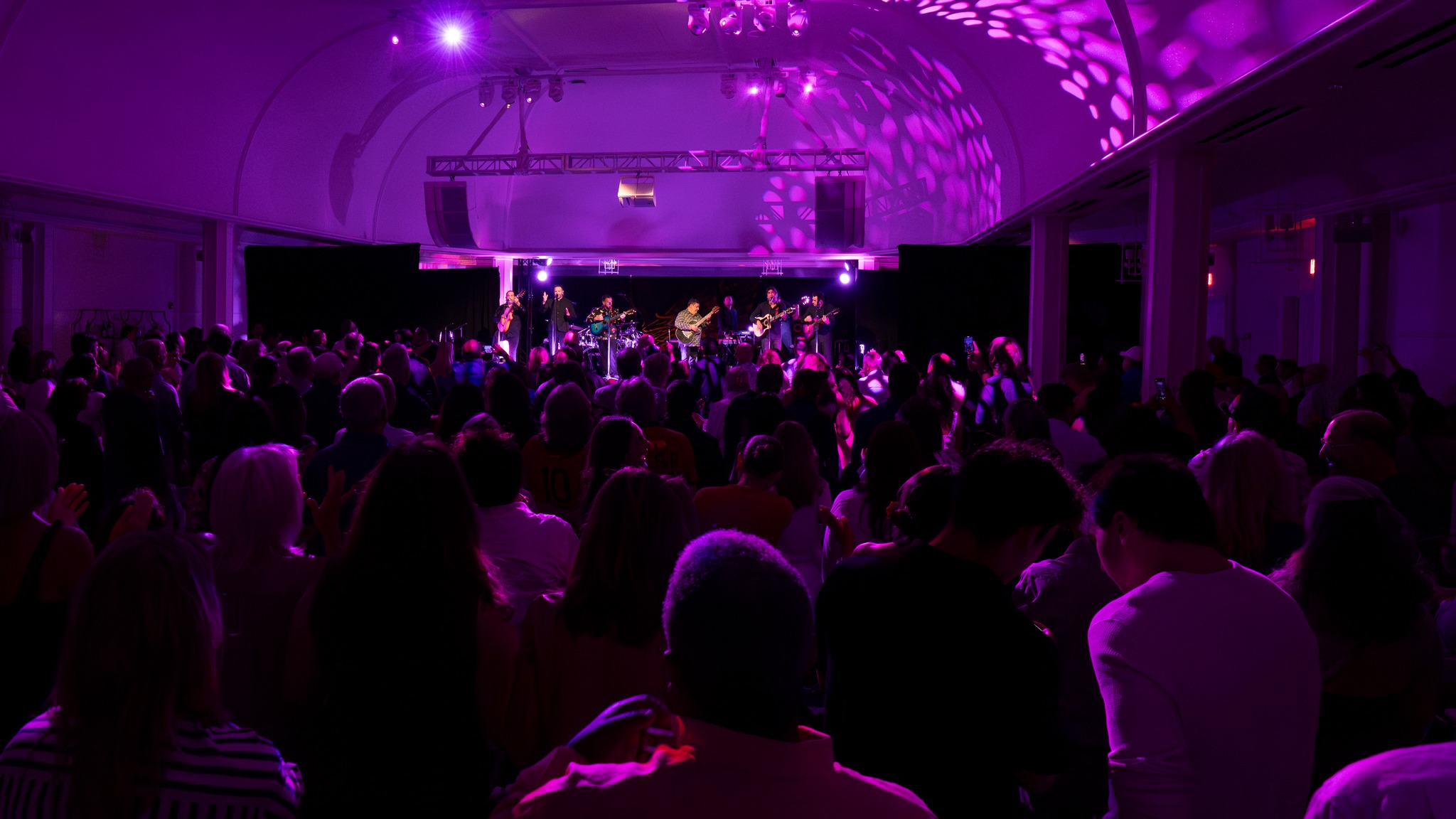 505584609_1021397906767606_8671512308669323074_n A large crowd watches a live band perform on stage under purple lighting in a spacious indoor venue.